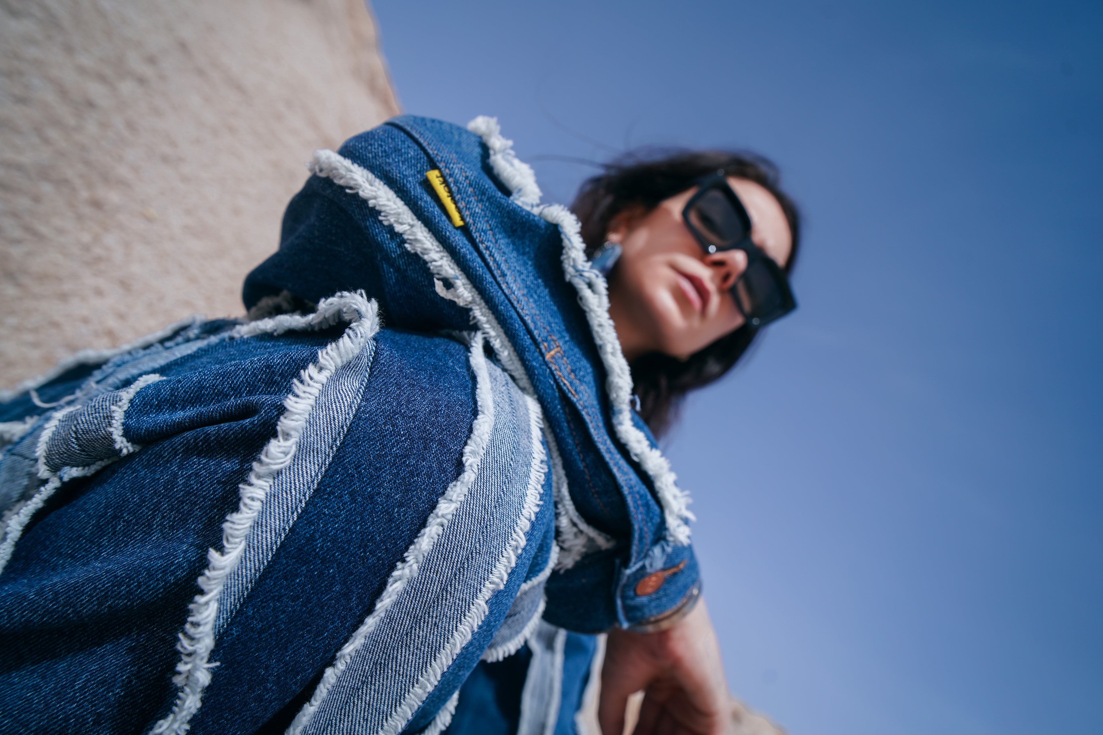 Mannequin portant une veste et un pantalon en denim upcyclé KONTAKT avec coutures effilochées visibles, photographiés en extérieur sous un ciel bleu
